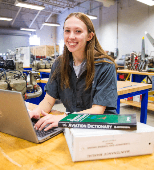 Female student sitting at workbench, with computer and aviation study guides, looking at camera, smiling.