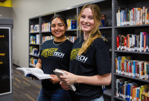 Two smiling female WSU Tech students standing next to each other in South campus library wearing WSU Tech black t-shirts with gold and white WSU Tech logos.