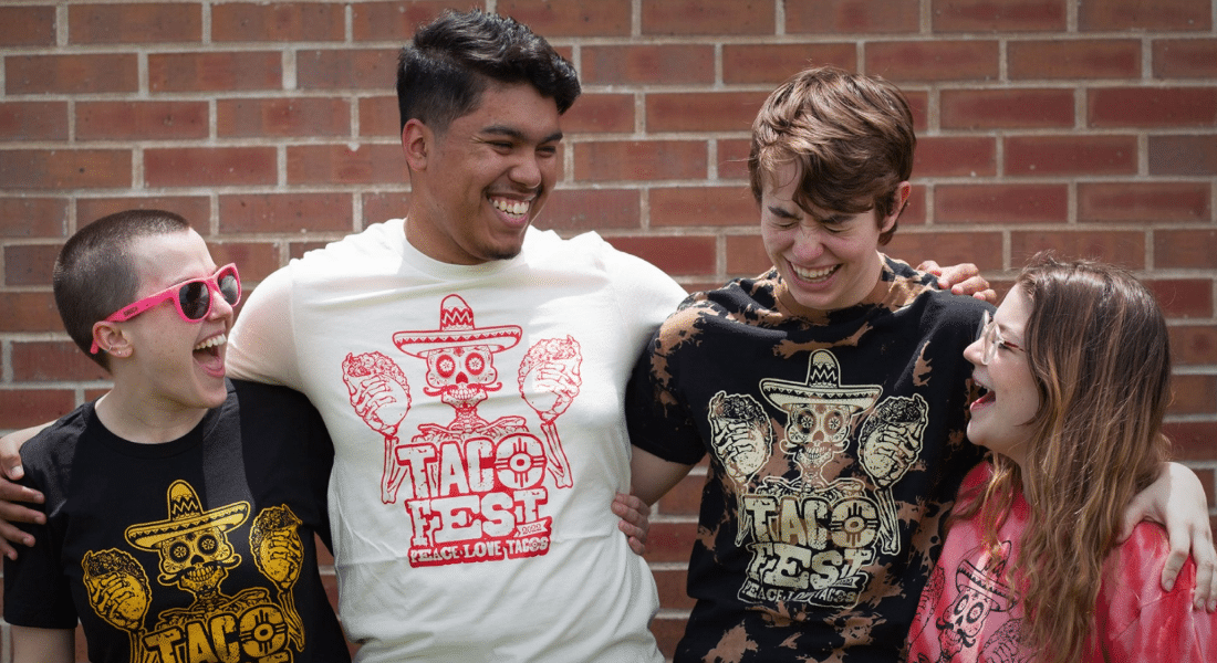 shock-starter-photography-main-3 Four friends wearing Taco Fest graphic t-shirts stand arm in arm, laughing together in front of a brick wall.