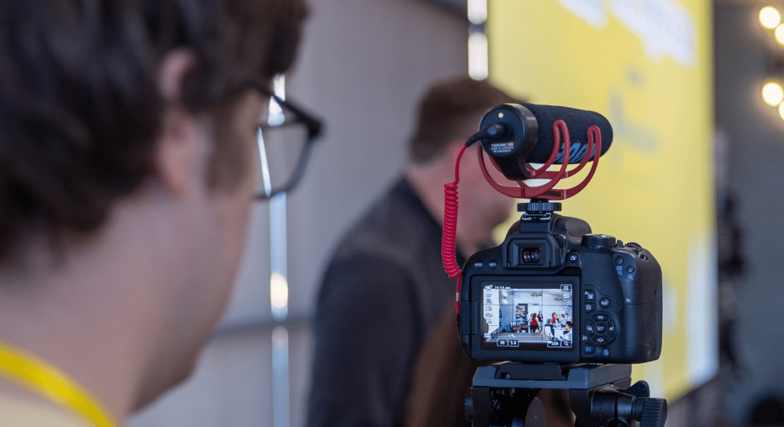 shock-starter-videography-main-5 Person filming with a DSLR camera on a tripod, capturing an interview in front of a yellow backdrop.