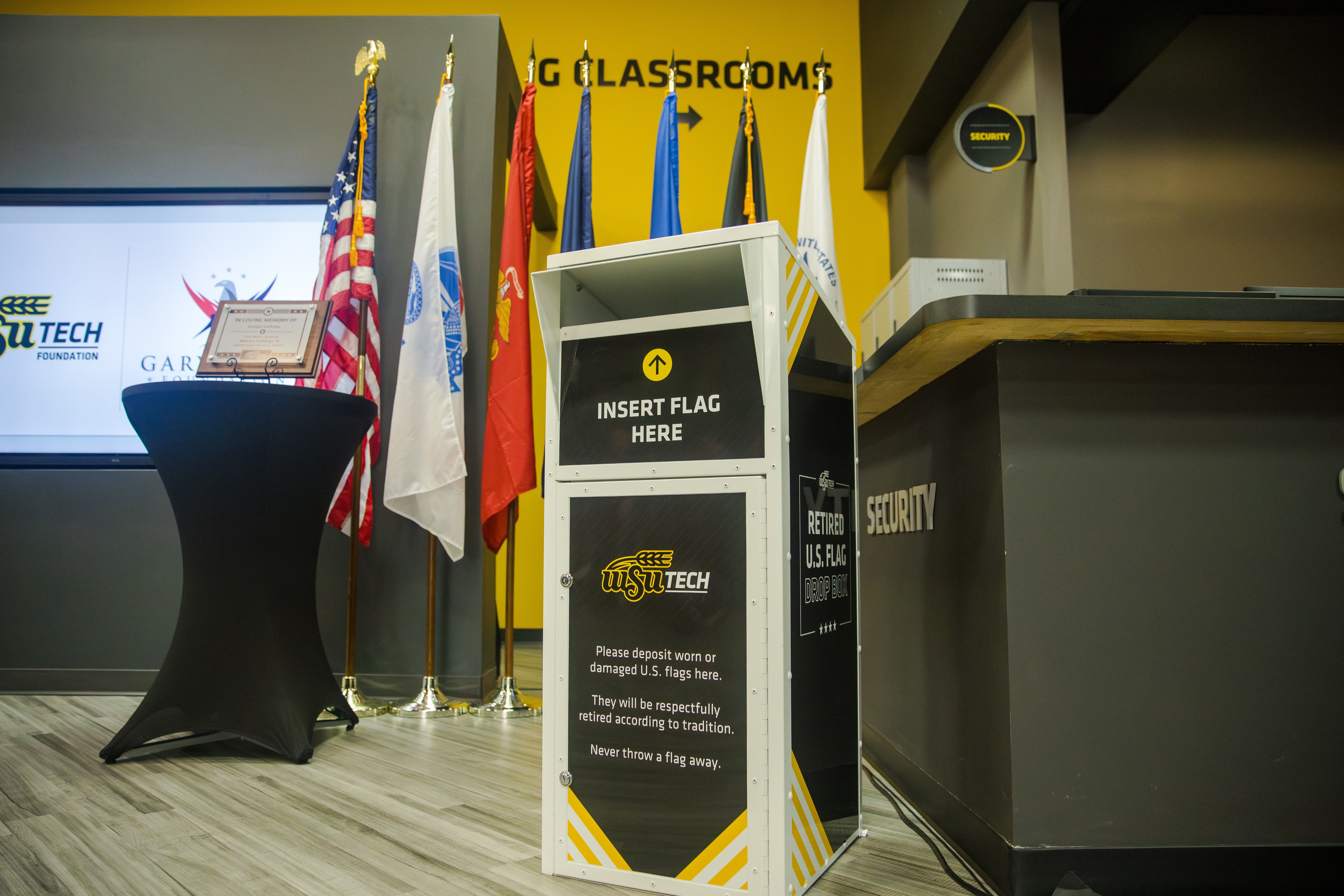 A flag deposit box with WSU TECH logo and DROP FLAG HERE on it stands in front of a flag display.
