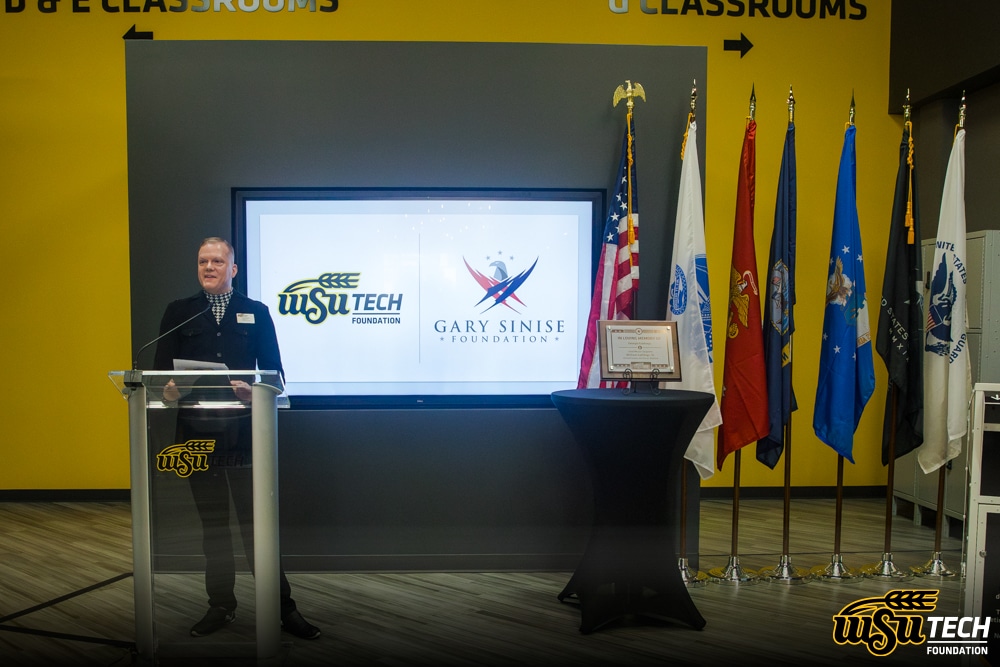 Photo of a man standing at a podium. WSU Tech Foundation logo on the podium. TV screen behind that has WSU Tech logo and Gary Sinese Foundation logo. Next to that is a flag display of the United States Flags and all US military branch flags. A plaque with the name Bill Gathings is on a table in front of the flags.