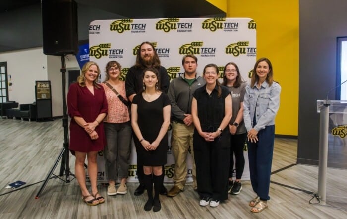 A group of 6 female adults and 2 male adults pose in front of a WSU Tech Foundation backdrop.