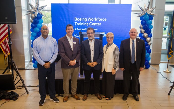 Five people pose in front of a stage display that reads “Boeing Workforce Training Center” during a Boeing and WSU Tech partnership announcement event. Blue, silver and white balloon columns frame the stage, with U.S. and state flags displayed nearby.