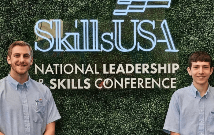 Skills USA National Leadership & Skills Conference sign on the back of a green leafy wall. Two young men smile.