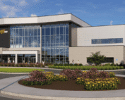 New National Center for Aviation Training building with large glass facade, landscaped circular flowerbeds and shrubs, parking area with cars, blue sky with clouds, and red “CROSSLAND TESSERE” text at the bottom right.