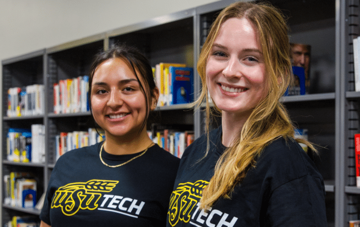 Two WSU Tech students smiling in front of bookshelves in the campus library.